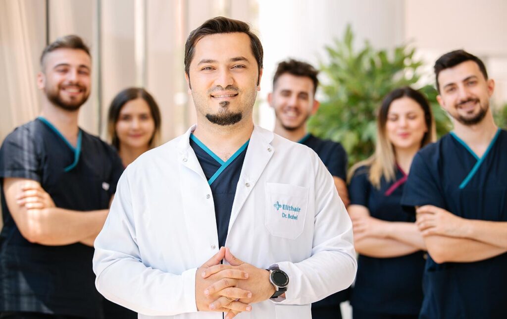 Dr. Balwi und sein medizinisches Team. Dr. Balwi steht im weißen Arztkittel im Vordergrund mit gefalteten Händen. Hinter ihm stehen fünf lächelnde Teammitglieder in dunkelblauen Kasacks. Alle blicken direkt in die Kamera.