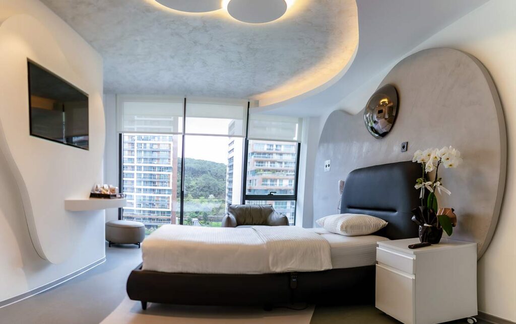 Ein luxuriöses Patientenzimmer in der Elithair Klinik. Ein gemachtes Bett mit weißer Bettwäsche, ein Flachbildfernseher an der Wand, eine geschwungene Deckenbeleuchtung, ein Nachttisch mit Orchideen und ein Sessel vor bodentiefen Fenstern, die Hochhäuser zeigen.