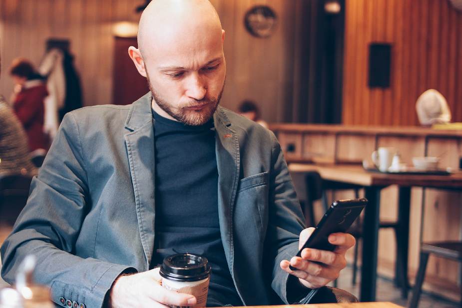 Hombre con calvicie total bebiendo café y mirando su teléfono
