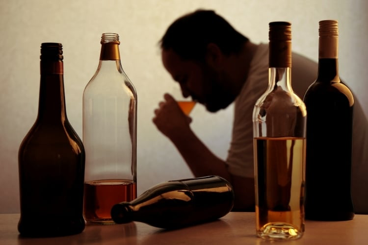 Diverse bottiglie davanti a un uomo dall'aria depressa che beve un bicchiere di alcolico