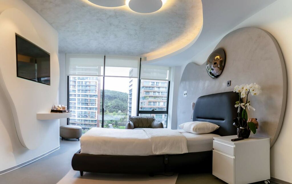 Ein luxuriöses Patientenzimmer in der Elithair Klinik. Ein gemachtes Bett mit weißer Bettwäsche, ein Flachbildfernseher an der Wand, eine geschwungene Deckenbeleuchtung, ein Nachttisch mit Orchideen und ein Sessel vor bodentiefen Fenstern, die Hochhäuser zeigen.