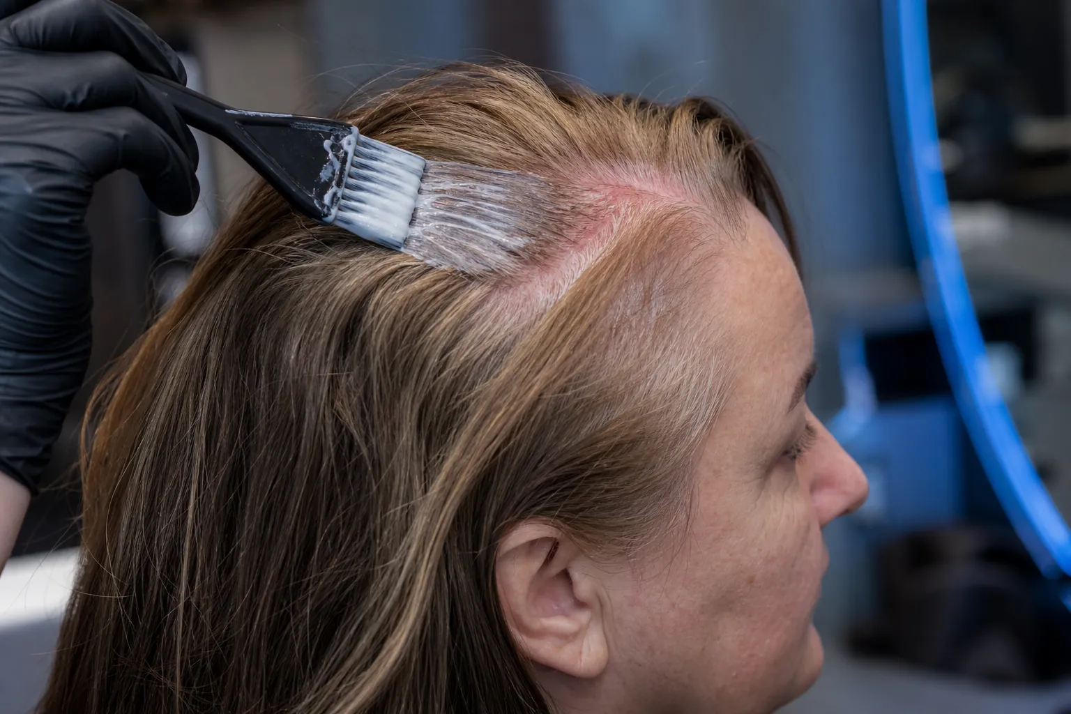 A hairdresser's hand brushes color cream along the part onto a reddened scalp