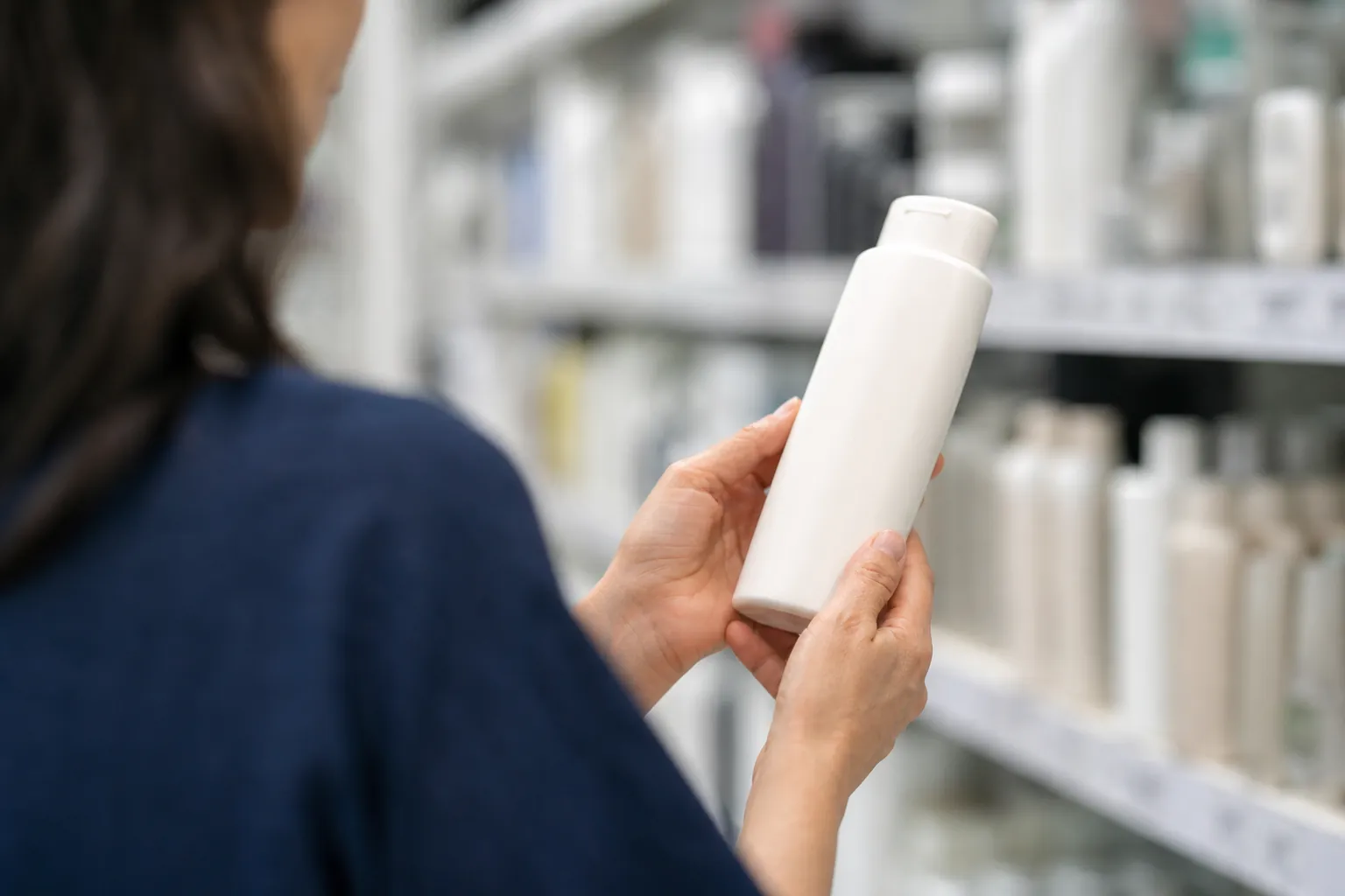 Woman in a drugstore aisle turning an unlabeled shampoo bottle around to read the ingredients